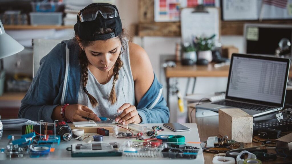 Real-World Application and Experience: Women with STEAM Phase 3 - Rock the STEAM Team Inc. A women in steam is gaining real-world application and experience working on circuitry with a computer in the background.